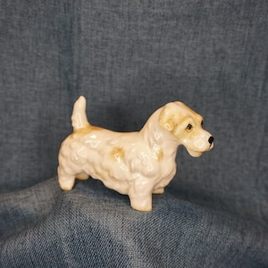 May include: A white ceramic figurine of a Scottish Terrier dog. The dog is standing with its head turned to the side and its tail curled. The figurine has brown markings on its body.