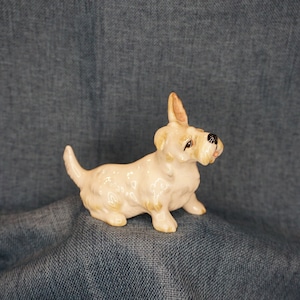 May include: A white ceramic figurine of a Scottish Terrier dog. The dog is sitting with its head turned to the side and its tongue sticking out.