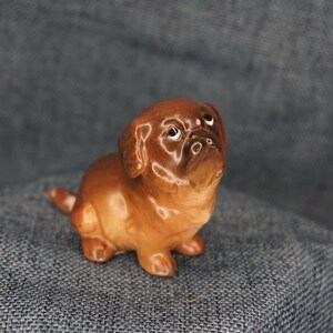 May include: A small brown ceramic figurine of a pug dog with a white nose and white markings around the eyes. The figurine is looking up.