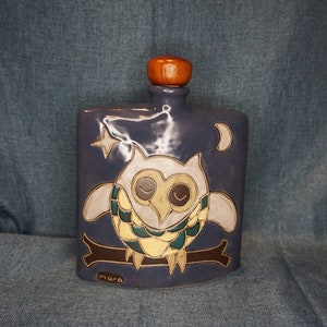 May include: A ceramic bottle with a wooden stopper, featuring a hand-painted owl with closed eyes, a crescent moon, and a star. The bottle is decorated with a blue, green, and yellow color scheme and has the word "mara" written on the bottom.