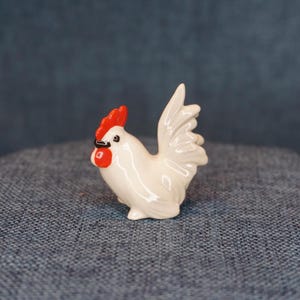 May include: A small, white ceramic rooster figurine with a bright red comb and wattle. The rooster has black accents around the eyes and beak. The figurine is posed in a standing position, with its tail feathers fanned out.