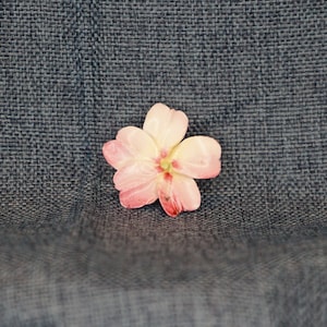 May include: A delicate pink flower with a white center, resembling a cherry blossom, is displayed on a textured gray fabric background.