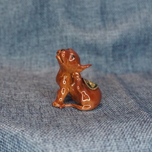 May include: A small, brown ceramic figurine of a dog with its head tilted up. The figurine has a glossy finish and is sitting on a blue denim fabric.