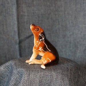 May include: A ceramic figurine of a brown and black dog with white markings. The dog is sitting up with its head tilted back and its mouth slightly open.