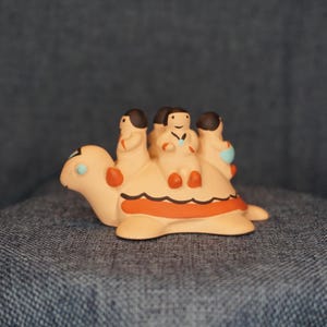 May include: A ceramic turtle figurine with four small figures riding on its back. The turtle is tan with orange and black accents, and the riders are wearing simple clothing. The figurine is set against a blue background.