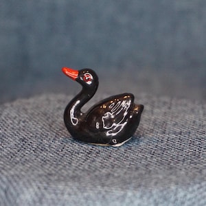 May include: A small, glossy black ceramic swan figurine with a red beak and red eyes. The swan is in a resting position, with its neck curved gracefully. The figurine is a decorative item.