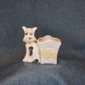 May include: A white ceramic planter with a dog design. The dog has yellow and black accents, with a small planter box attached. The planter box has a design of small yellow dogs.