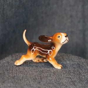 May include: A small ceramic figurine of a beagle puppy in a running pose. The dog has a glossy finish with brown, tan, and white markings, large floppy ears, a raised tail, and painted black eyes and nose, capturing a playful and active stance.