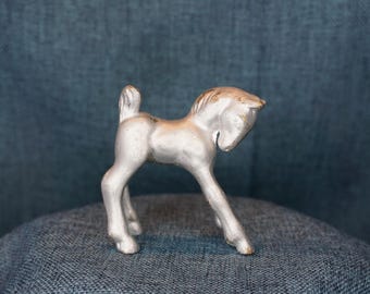 May include: A silver metal figurine of a horse with a raised tail. The horse is standing on a blue fabric surface.