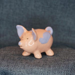 May include: A small pink ceramic pig figurine with purple heart-shaped markings. The pig has a cute, cartoon-like face and is standing on all four legs.