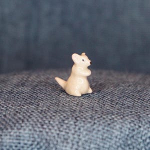 May include: A small, white ceramic figurine of a kangaroo standing on a blue fabric surface.
