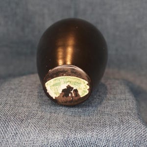 May include: A small, dark brown, egg-shaped vase with a glossy finish. The base has a partially visible, faded sticker with text. The vase is likely vintage or antique.