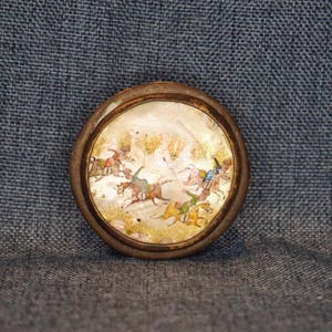 May include: A circular brooch featuring a polished mother-of-pearl inlay set within a dark metallic frame. The surface displays a detailed hand-painted scene of multiple figures riding horses across a light, textured background with gold accents.