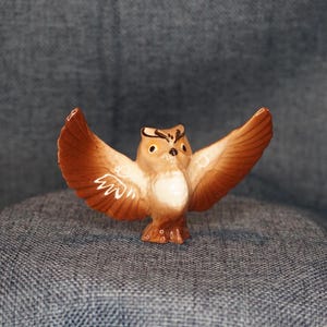 May include: A small ceramic owl figurine with brown and white painted details. The owl has its wings spread out, as if it is flying.