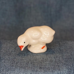 May include: A small, white ceramic chick figurine with a red beak and accents. The chick is in a downward-facing pose, with a small black dot for an eye. The figurine is on a blue denim surface.