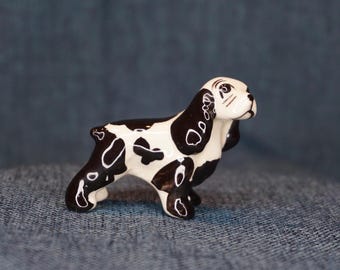 May include: A ceramic figurine of a dog, primarily white with black spots and markings. The dog is in a standing pose, with its head turned slightly to the side. The figurine has detailed features, including the eyes, ears, and paws. The background is a solid blue.