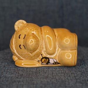 May include: A ceramic figurine of a snail with a cartoonish design. The snail is light brown with black details and a small bee illustration. The figurine has a glossy finish and is resting on a blue surface.
