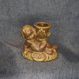 May include: A brown ceramic candle holder featuring a cherub figure holding a decorative candle cup. The cherub is sitting on a base with intricate floral designs.
