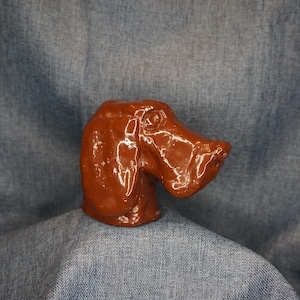 May include: A brown ceramic dog head figurine with a glossy finish. The dog's head is sculpted with detailed features, including a long snout, visible eye, and textured fur. The figurine is a decorative item, likely for display.