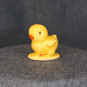 May include: A small, yellow ceramic chick figurine with an orange beak and black eye. The chick is standing on a small, round base. The figurine is a decorative item.