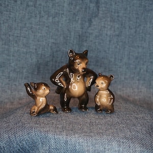 May include: A set of three glossy brown ceramic bear figurines. One larger bear stands in the center with hands on hips, while two smaller bears are positioned on either side, one sitting and one standing, all featuring smooth, reflective surfaces.