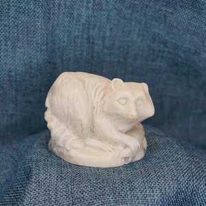 May include: A white ceramic figurine of a raccoon sitting on its haunches. The figurine is detailed with fur and facial features.