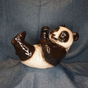 May include: A ceramic panda figurine with black and white fur, lying on its back with its legs in the air. The panda has a glossy finish and realistic details.