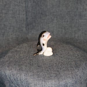 May include: A small ceramic dolphin figurine. The dolphin is primarily white with black and gray accents. It has a pink mouth and cheeks, and a small, white base. The figurine is posed as if leaping or jumping.