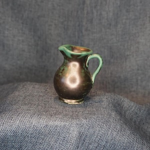 May include: A small, handcrafted ceramic pitcher with a dark brown body and a green rim and handle. The pitcher has a rounded shape and a small spout. The glaze has a mottled appearance, adding to its rustic charm. The pitcher is likely used for serving liquids.