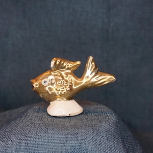 May include: A small gold ceramic fish figurine with a white base. The fish has a detailed design and a single eye.