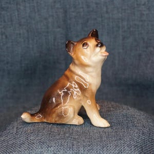 May include: A ceramic figurine of a dog in a sitting position, with its head tilted upwards and tongue sticking out. The dog has brown and tan fur with white accents. The figurine is a decorative item.