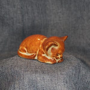 May include: A small, ceramic figurine of a sleeping cat. The cat is a warm, caramel brown color with white accents on its paws and belly. The cat is curled up, with its head resting on its paws. A charming decorative item.