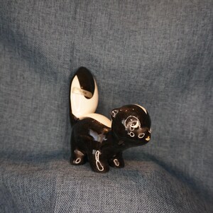 May include: A black and white ceramic figurine of a skunk with a white stripe down its back and a black tail. The skunk has a cute, cartoon-like face with black eyes and a small, pink nose.