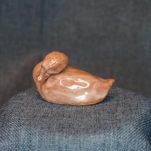 May include: A ceramic duck figurine in shades of brown, with a glossy finish. The duck is in a resting pose, with its head tucked down. The figurine is placed on a textured, blue-gray surface.