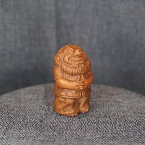 May include: A small, wooden figurine of a smiling, bearded man with his hands clasped. The figure is carved in a detailed style, with visible clothing and facial features. The wood has a warm, natural tone.