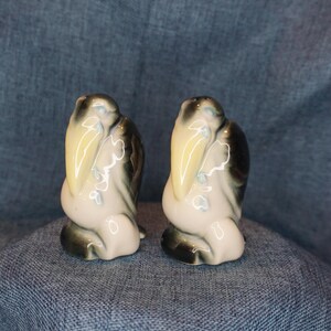May include: A pair of ceramic salt and pepper shakers shaped like pelicans. The pelicans are white with black and yellow accents. They are sitting on a blue fabric surface.