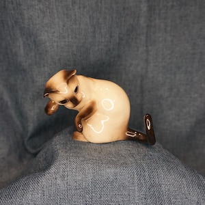 May include: A ceramic Siamese cat figurine with a cream-colored body and dark brown points on the ears, face, and tail. The cat is depicted in a seated position with one paw raised toward its head, featuring a glossy finish and smooth curves.