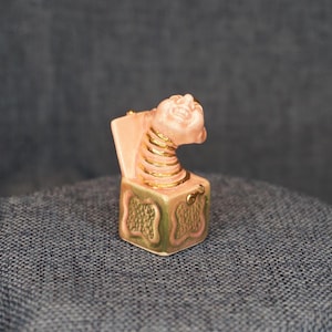 May include: A miniature jack-in-the-box figurine featuring a pink, smiling face attached to a coiled spring neck. The square base is painted in shades of pink and gold with a textured, embossed pattern on the sides and a small gold crank handle.
