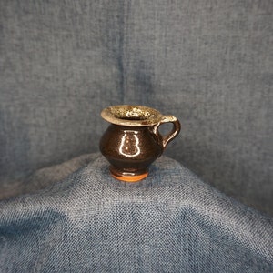 May include: A small brown ceramic pitcher with a light brown rim and a handle. The pitcher is sitting on a blue fabric surface.