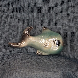 May include: A ceramic fish figurine featuring a glossy finish with blended shades of teal, blue, and brown. The fish has a curved tail, defined fins, and a large, dark eye with painted eyelashes, presented in a side profile view with a smooth texture.