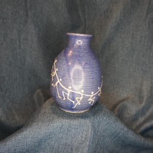 May include: A small, blue ceramic vase with a rounded body and a narrow neck. The vase features a white, stylized design of figures holding hands around its circumference. The vase is approximately 6 inches tall.