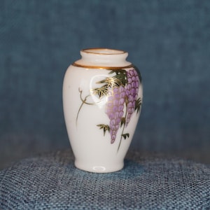 May include: Small white ceramic vase with a gold rim and a purple wisteria design. The vase features a detailed illustration of wisteria flowers and leaves, with gold accents. The vase is a decorative item.