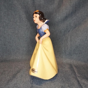 May include: A ceramic figurine of Snow White from Disney's Snow White and the Seven Dwarfs. She is wearing a yellow dress with a blue bodice and a red bow in her hair. The figurine is standing on a blue background.
