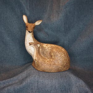 May include: A ceramic sculpture of a deer with a fawn nestled against its side. The deer has a textured brown and white finish, with a long neck, upright ears, and a smooth, rounded body shape. The fawn is smaller, mirroring the deer's colors.