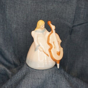 May include: A porcelain figurine of a girl playing a cello. The girl is wearing a white dress with blue accents and has blonde hair. The cello is brown with gold accents.