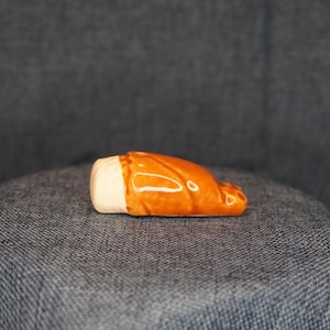 May include: A small, ceramic beer stein figurine. The stein is primarily orange with a white base and a handle. The figurine is lying on its side, showcasing its detailed design and glossy finish. The background is a textured, dark gray surface.