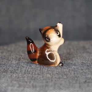 May include: A small ceramic raccoon figurine with a brown and white body, black stripes on its tail, and a curious upward gaze. The figurine has a glossy finish and is set against a neutral background.