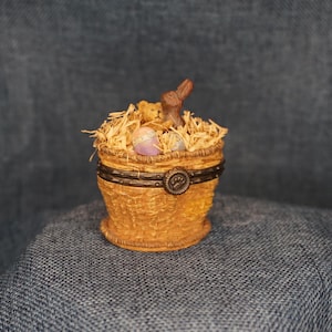 May include: A round, tan, woven-textured trinket box featuring a dark brown band with a central paw print emblem. The lid is filled with light-colored straw, a small brown teddy bear, a brown rabbit figurine, and two pastel-colored eggs.