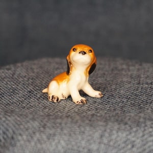 May include: A small ceramic figurine of a beagle puppy. The puppy is sitting with its front paws extended. The figurine has a white body with brown and black markings on its head and ears. The puppy's eyes are black.