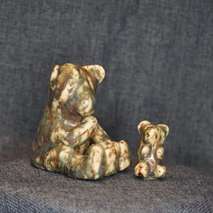 May include: Two ceramic bear figurines with a glossy, mottled glaze featuring blended shades of earthy brown, sage green, and cream. The larger bear sits in a rounded posture, while the smaller bear stands upright beside it, both smooth.